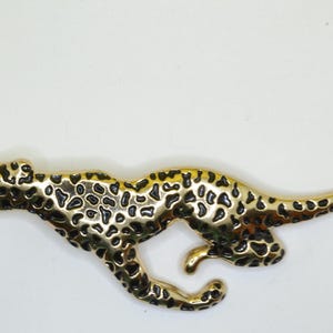 Puede incluir: Figura de guepardo de metal dorado con manchas negras. El guepardo está representado en una pose de carrera, con el cuerpo alargado y la cola curvada. Es probable que la figura sea un objeto decorativo.