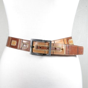 Op de afbeelding: Een bruine leren riem met een zilveren gesp. De riem heeft een geometrisch patroon van vierkanten en rechthoeken in bruin en beige.