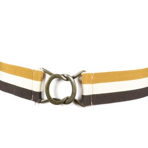 Könnte beinhalten: Ein gestreifter Gürtel mit einer bronzefarbenen Metallschnalle. Der Gürtel hat braune, weiße und goldfarbene Streifen. Die Schnalle hat ein kreisförmiges Design mit Knotenoptik. Der Gürtel wird auf einer weißen Form präsentiert.