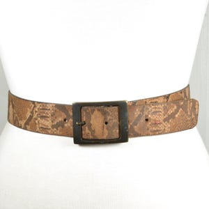 Könnte beinhalten: Ein brauner Gürtel mit Schlangenmuster und einer quadratischen, dunklen Metallschnalle. Der Gürtel ist um eine weiße Form gewickelt und zeigt sein Design und seine Textur. Die Gürtelbreite beträgt ca. 3,8 cm.