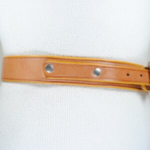 A tan leather belt with metal studs and a buckle is shown on a mannequin.