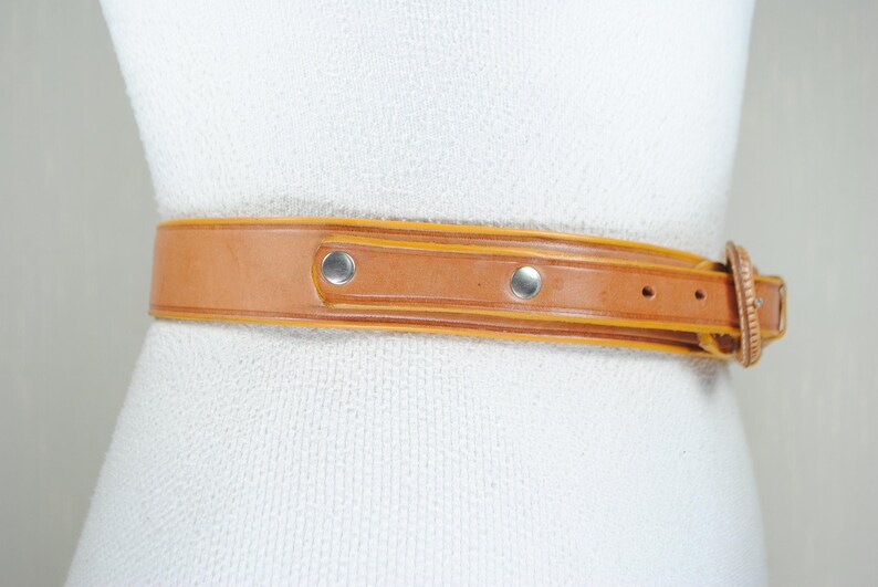 A tan leather belt with metal studs and a buckle is shown on a mannequin.