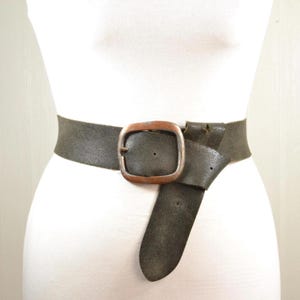 May include: A wide, dark olive green belt featuring a textured surface and a large, rounded rectangular buckle with a metallic copper finish. The belt strap passes through the buckle and hangs downward, displaying a single prong and visible holes.