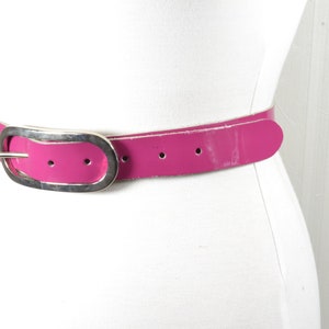 Könnte beinhalten: Ein leuchtend pinkfarbener Gürtel mit einer silberfarbenen Schnalle. Der Gürtel besteht aus glänzendem Material und wird auf einer weißen Schaufensterpuppe präsentiert. Die Schnalle hat eine ovale Form.