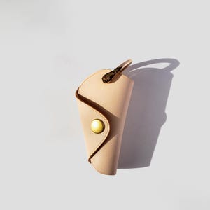 May include: A light beige leather key holder with a brass button and a metal ring. The key holder is designed to hold keys securely.