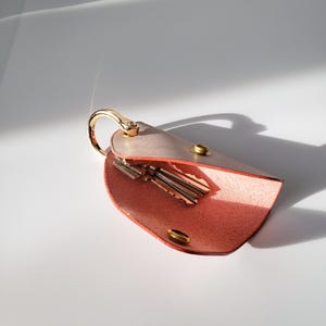 May include: A pink leather key holder with a gold metal ring and a flap that opens to reveal a space for keys. The holder is open and shows three keys inside.