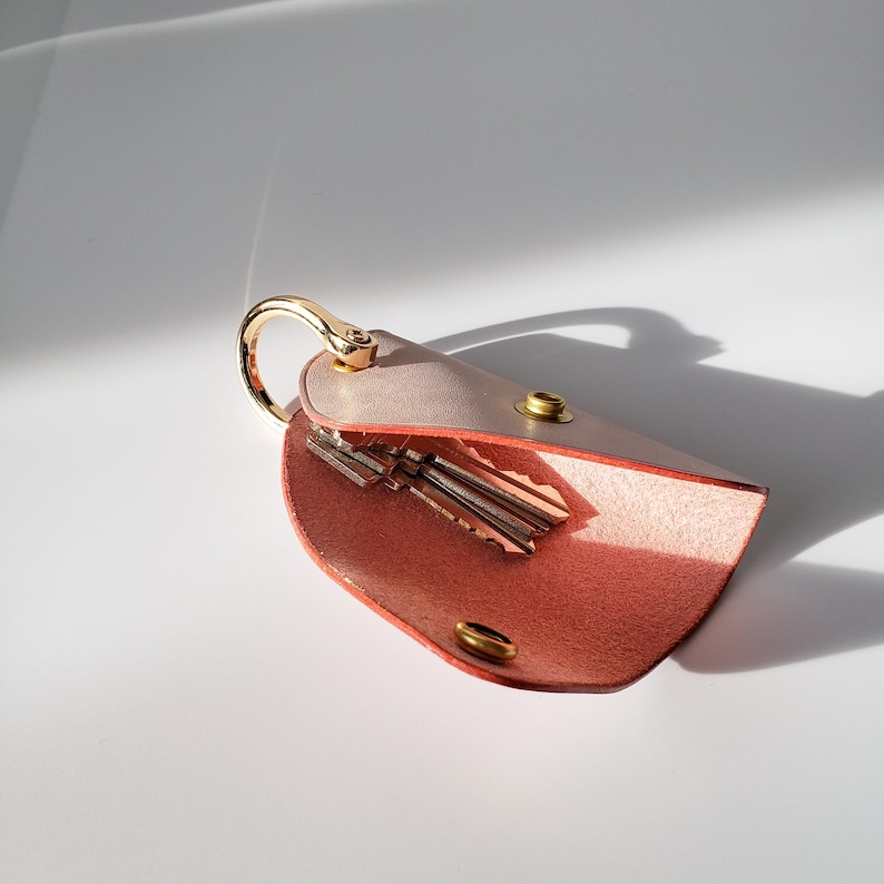 May include: A pink leather key holder with a gold metal ring and a flap that opens to reveal a space for keys. The holder is open and shows three keys inside.