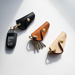 May include: Three leather key holders in black, brown, and beige, each with a brass snap closure and a brass key ring. One holder is attached to a car key fob. The key holders are arranged on a white surface.