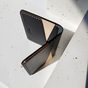 Puede incluir: Cartera de cuero negro con un acento beige y costuras blancas. La cartera tiene un dise&ntilde;o minimalista con la palabra "BAKUL" en relieve en la parte delantera. Los bordes est&aacute;n acabados con un ribete marr&oacute;n.