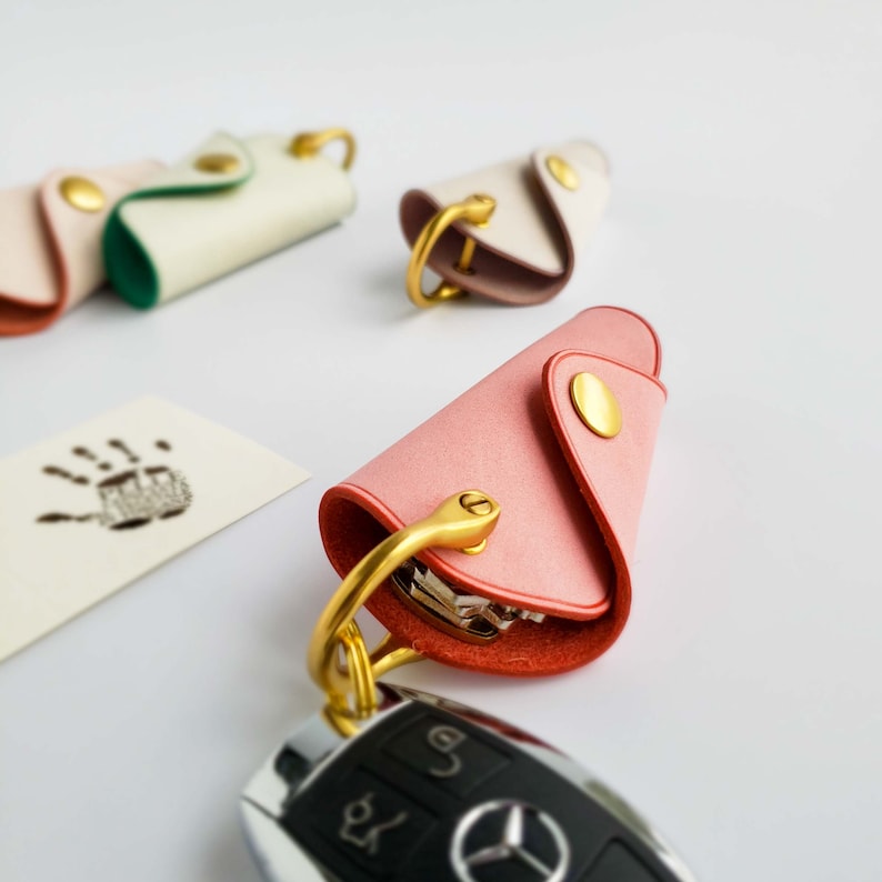 May include: A set of four leather keychains in pink, white, and green. The keychains are shaped like a folded flap and have a gold metal ring and clasp. The keychains are attached to a silver car key.