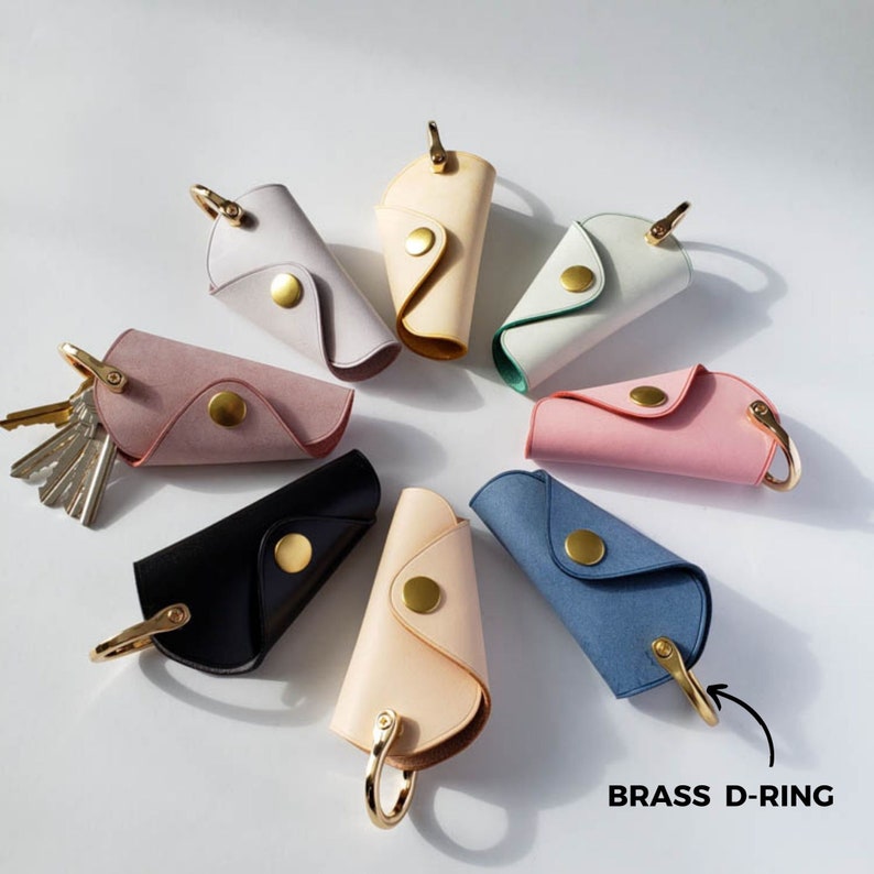 Puede incluir: Un conjunto de ocho llaveros de cuero en varios colores, incluyendo rosa, azul, beige y negro. Cada llavero tiene un anillo en D de lat&oacute;n y un dise&ntilde;o plegable.