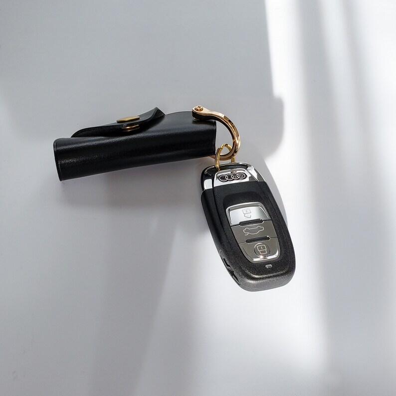 Puede incluir: Un llavero de cuero negro con un anillo de llave dorado y un llavero de coche negro con botones plateados. El llavero tiene la palabra "Audi" impresa en &eacute;l.