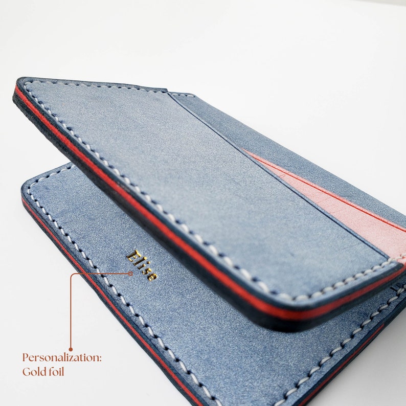 Puede incluir: Cartera de cuero azul con detalles rojos y blancos. La cartera presenta una franja roja y costuras blancas. La palabra "Elise" est&aacute; grabada en pan de oro. Un accesorio elegante.