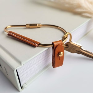 May include: A gold key ring with a brown leather strap and a small gold button. The key ring is attached to a book.