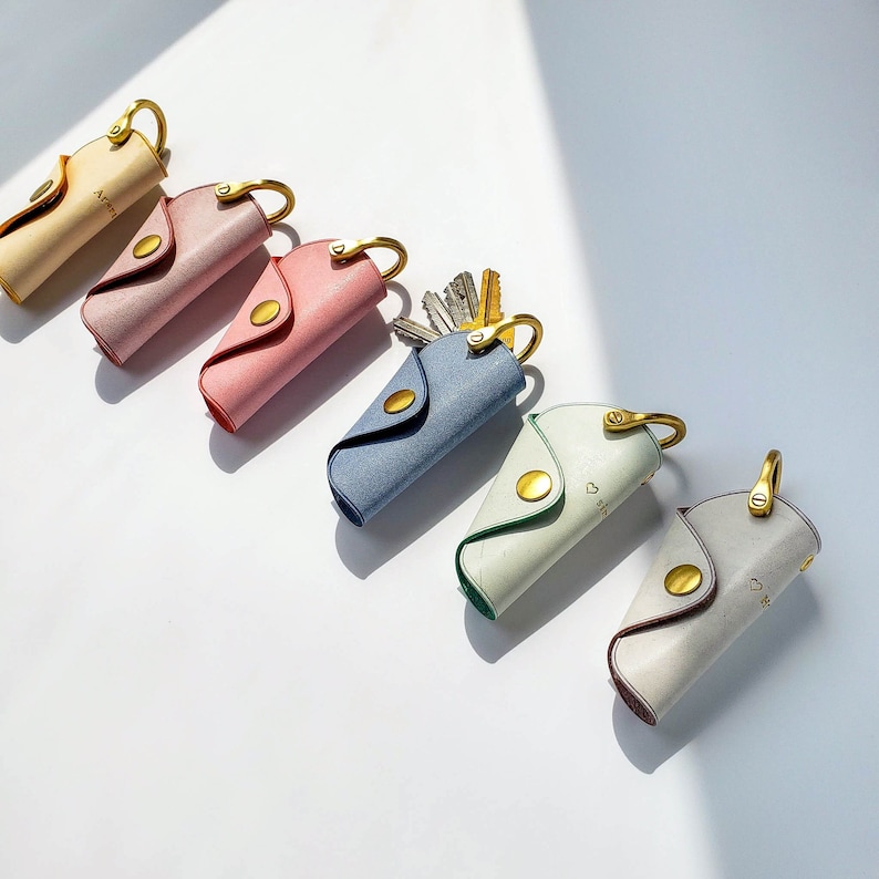 May include: Six leather keychains in various colors, including pink, blue, and white. Each keychain has a gold metal ring and a small metal charm. The keychains are arranged in a row on a white surface.