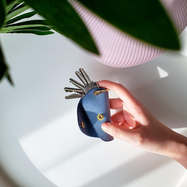 May include: A blue leather key holder with a flap that opens to reveal a set of keys. The holder has a gold snap closure and is held in a hand.