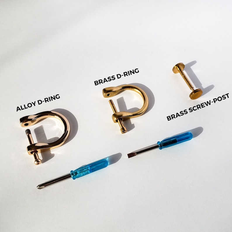 Puede incluir: Dos anillos en D de color dorado, uno etiquetado como "Brass D-Ring" y el otro como "Alloy D-Ring", se muestran junto a un poste de tornillo de lat&oacute;n etiquetado como "Brass Screw-Post". Tambi&eacute;n se muestran dos destornilladores azules.