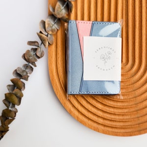 Puede incluir: Una peque&ntilde;a cartera bicolor en un envoltorio de pl&aacute;stico transparente. La cartera es azul claro y rosa con una etiqueta blanca que dice "SERENIKEY HANDMADE" en un dise&ntilde;o circular. La cartera est&aacute; sobre una bandeja de madera semicircular.
