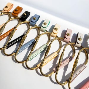 Puede incluir: Un conjunto de diez llaveros con correas de cuero de diferentes colores. Cada llavero está hecho de latón y tiene una forma ovalada única. Las correas están cosidas con hilo blanco.