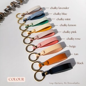 May include: A set of ten leather keychains in various colours, including chalky lavender, chalky blue, chalky mint, chalky lemon, chalky pink, chalky rose, beige, tan, and black. The keychains are arranged in a row from lightest to darkest. The text "COLOUR" is at the bottom left of the image. The text "Loop Keychains, No Personalization" is at the bottom right of the image.