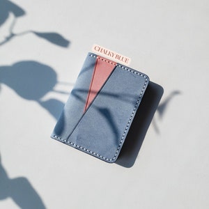 Puede incluir: Un tarjetero de cuero azul tiza con un acento rosa diagonal. El tarjetero est&aacute; cosido con hilo blanco en los bordes. El texto "CHALKY BLUE" est&aacute; impreso en una peque&ntilde;a etiqueta.