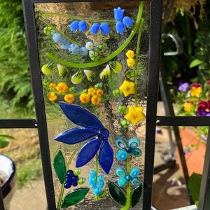 May include: A rectangular stained glass panel with a black metal frame. The panel features a colorful floral design with blue, yellow, and green glass elements. The design includes bluebell-like flowers, a large blue flower, and other stylized floral shapes.