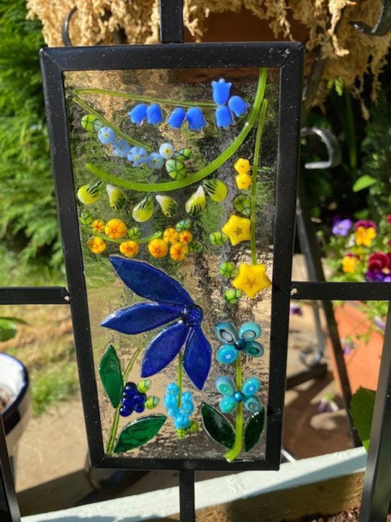 May include: A rectangular stained glass panel with a black metal frame. The panel features a colorful floral design with blue, yellow, and green glass elements. The design includes bluebell-like flowers, a large blue flower, and other stylized floral shapes.