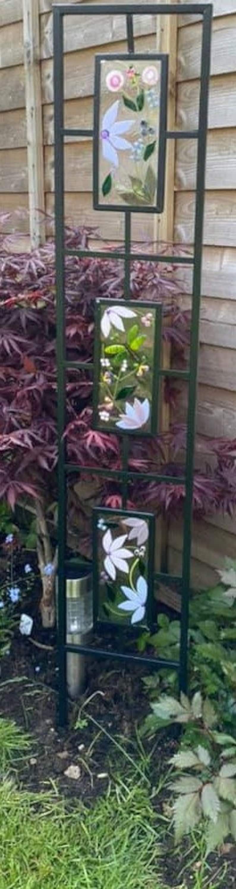 May include: A black metal garden trellis with three rectangular glass panels. Each panel features a floral design with white, pink, and blue flowers. The trellis is set in a garden with green grass and purple foliage.
