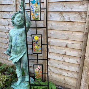 May include: A black metal garden trellis with three rectangular glass panels. Each panel features a colorful floral design, including a blue butterfly, yellow sun, and pink flower. The trellis is placed in a garden setting with a statue.