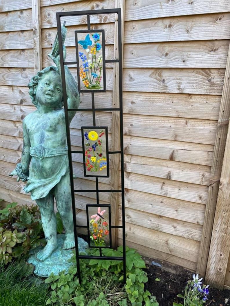 May include: A black metal garden trellis with three rectangular glass panels. Each panel features a colorful floral design, including a blue butterfly, yellow sun, and pink flower. The trellis is placed in a garden setting with a statue.