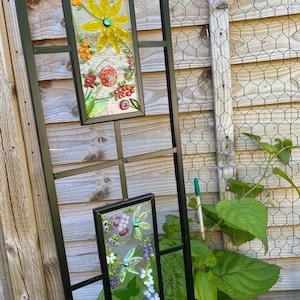 May include: Two rectangular stained glass panels with floral designs hang on a black metal trellis. The top panel features a yellow flower and orange berries. The bottom panel displays purple grapes and white flowers.
