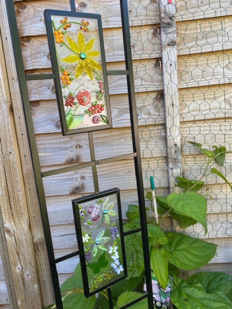 May include: Two rectangular stained glass panels with floral designs hang on a black metal trellis. The top panel features a yellow flower and orange berries. The bottom panel displays purple grapes and white flowers.