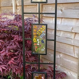May include: A black metal garden trellis with three rectangular panels, each displaying colorful floral and butterfly mosaic art. The trellis is set against a wooden fence and lush greenery.