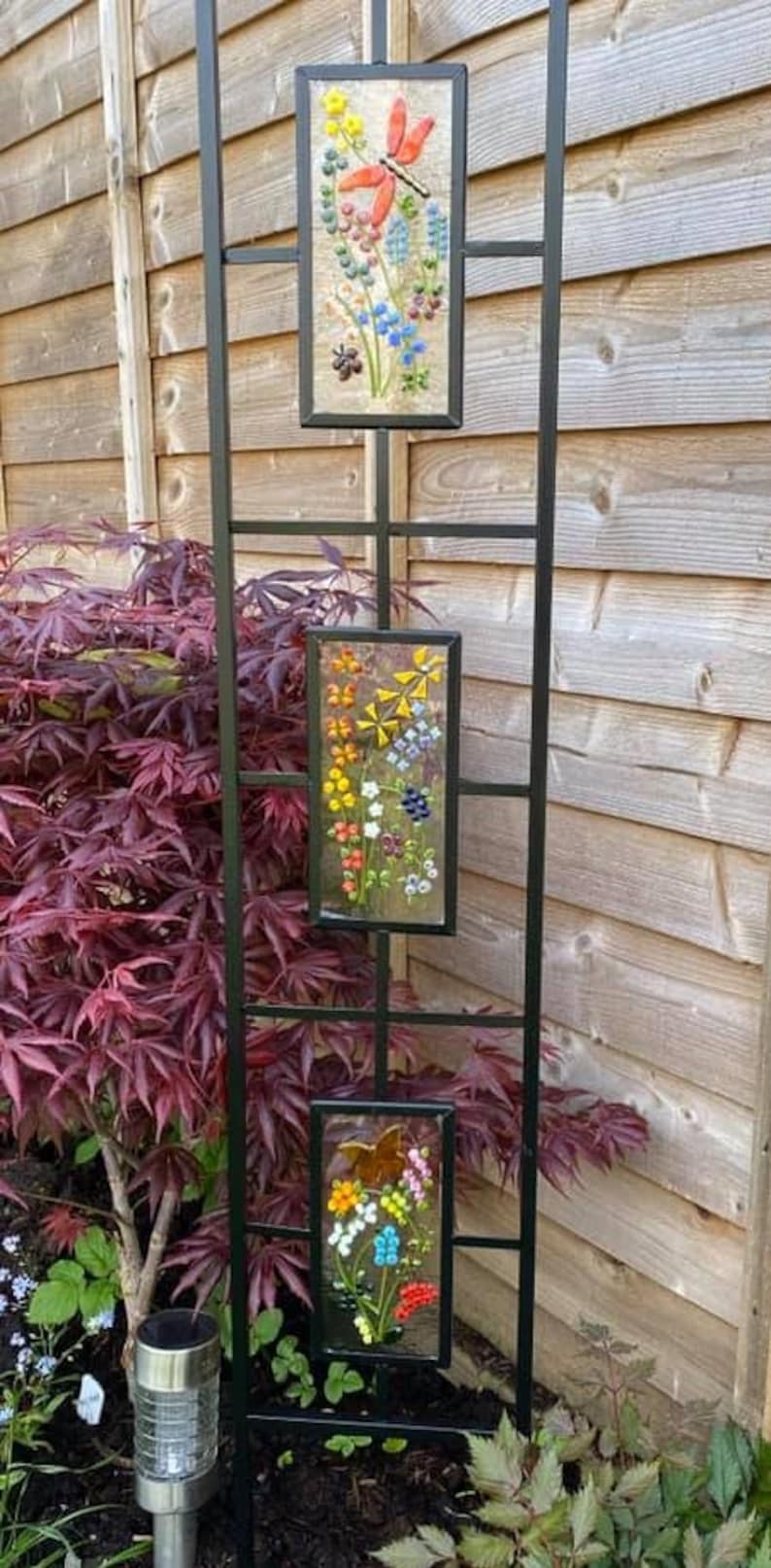 May include: A black metal garden trellis with three rectangular panels, each displaying colorful floral and butterfly mosaic art. The trellis is set against a wooden fence and lush greenery.