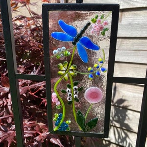 May include: A rectangular, stained-glass panel featuring a blue dragonfly and colorful floral elements. The artwork is set within a black metal frame, suitable for garden decor. The dragonfly has blue wings and a black body, with pink and yellow flowers.