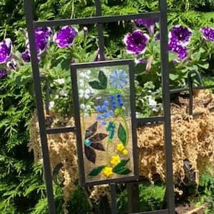 May include: A decorative metal garden trellis with three rectangular glass panels. Each panel features a colorful floral design with blue, yellow, and white flowers, and a butterfly. The trellis is set against a backdrop of green foliage and purple flowers.