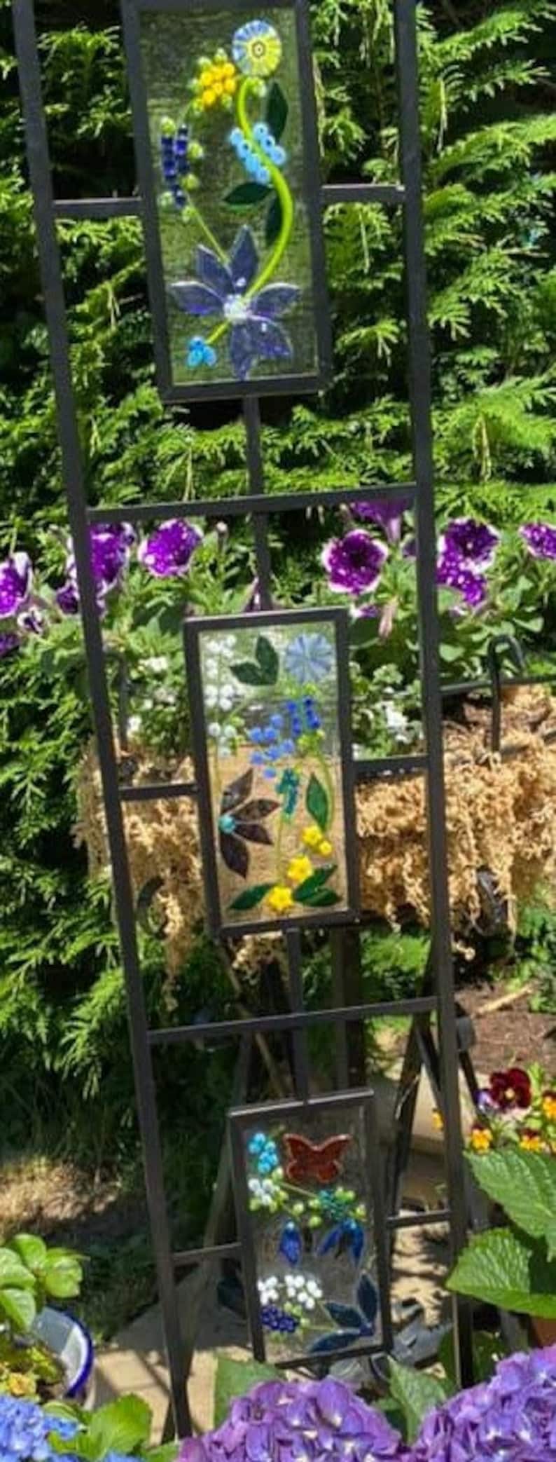 May include: A decorative metal garden trellis with three rectangular glass panels. Each panel features a colorful floral design with blue, yellow, and white flowers, and a butterfly. The trellis is set against a backdrop of green foliage and purple flowers.