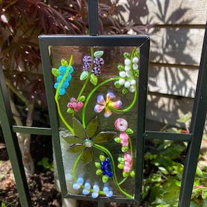 May include: A decorative garden stake featuring a rectangular glass panel with a floral design. The panel has a black frame and is adorned with colorful glass flowers, leaves, and berries in shades of blue, pink, purple, and green. The stake is made of black metal.
