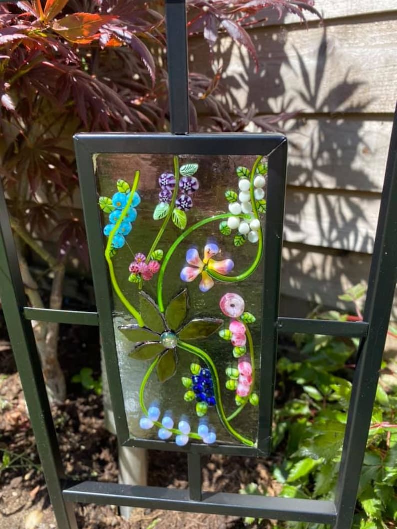 May include: A decorative garden stake featuring a rectangular glass panel with a floral design. The panel has a black frame and is adorned with colorful glass flowers, leaves, and berries in shades of blue, pink, purple, and green. The stake is made of black metal.