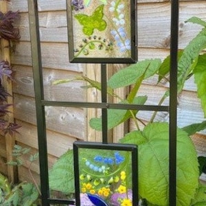 May include: Black metal garden trellis with two rectangular stained glass panels. The panels feature floral designs with butterflies, flowers, and leaves in shades of yellow, blue, purple, and green. The trellis is set against a wooden fence.