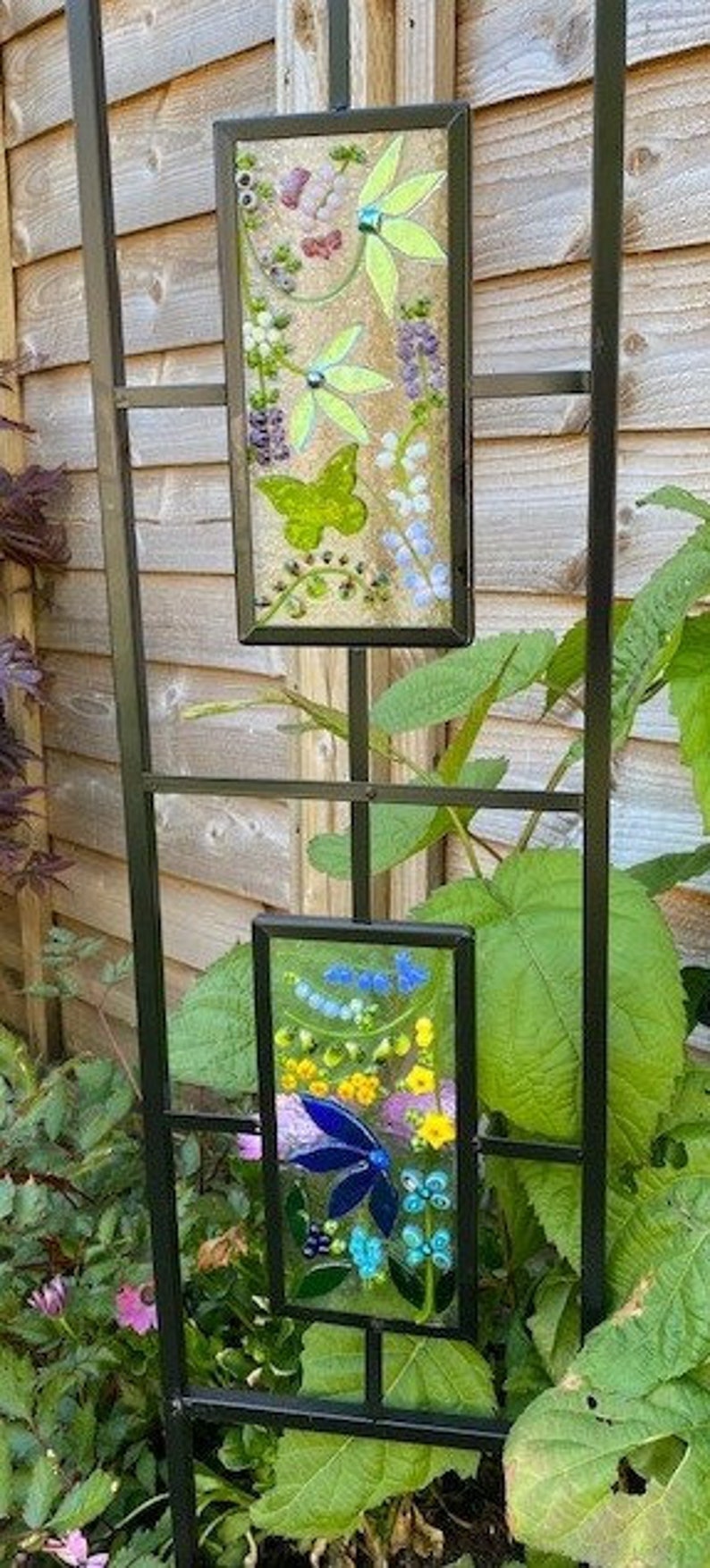 May include: Black metal garden trellis with two rectangular stained glass panels. The panels feature floral designs with butterflies, flowers, and leaves in shades of yellow, blue, purple, and green. The trellis is set against a wooden fence.
