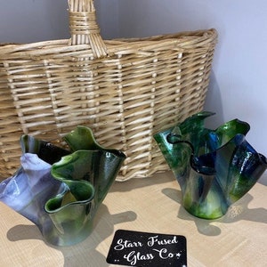 May include: Two handcrafted glass bowls with ruffled edges in shades of green, blue, and purple sit on a wooden surface. A woven basket is in the background. A black sign with white text reads "Starr Fused Glass Co."