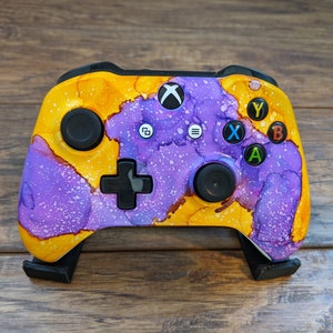 May include: A purple and yellow Xbox controller with a custom paint job. The controller has a black faceplate with the standard Xbox buttons and a black d-pad.