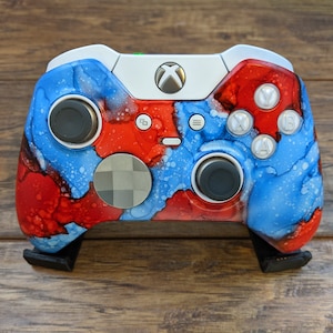 May include: A custom Xbox controller with a white base and a red, blue, and white abstract paint design. The controller has black buttons and a black D-pad.