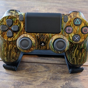 May include: A camouflage-patterned PlayStation 4 controller with black buttons and a black speaker grille. The controller is on a black stand.