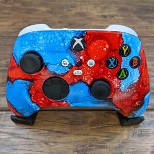 May include: A custom Xbox controller with a blue and red abstract paint design. The controller has black buttons and a white Xbox logo.