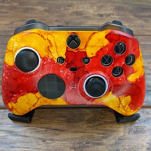 May include: A custom Xbox controller with a red and yellow abstract paint design. The controller has black buttons and a black D-pad.