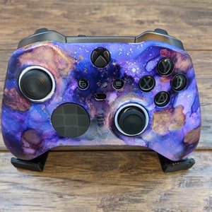 May include: A black Xbox controller with a custom purple and gold galaxy design. The controller has white accents around the buttons and joysticks.