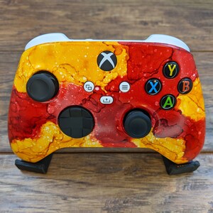Pode incluir: Um controlador Xbox personalizado com um design abstrato vermelho, amarelo e laranja. O controlador tem botões pretos e um D-pad preto.