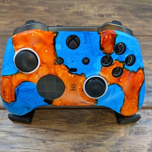 May include: A blue and orange Xbox controller with custom paint job. The controller has black buttons and a black d-pad. The controller is sitting on a wooden surface.